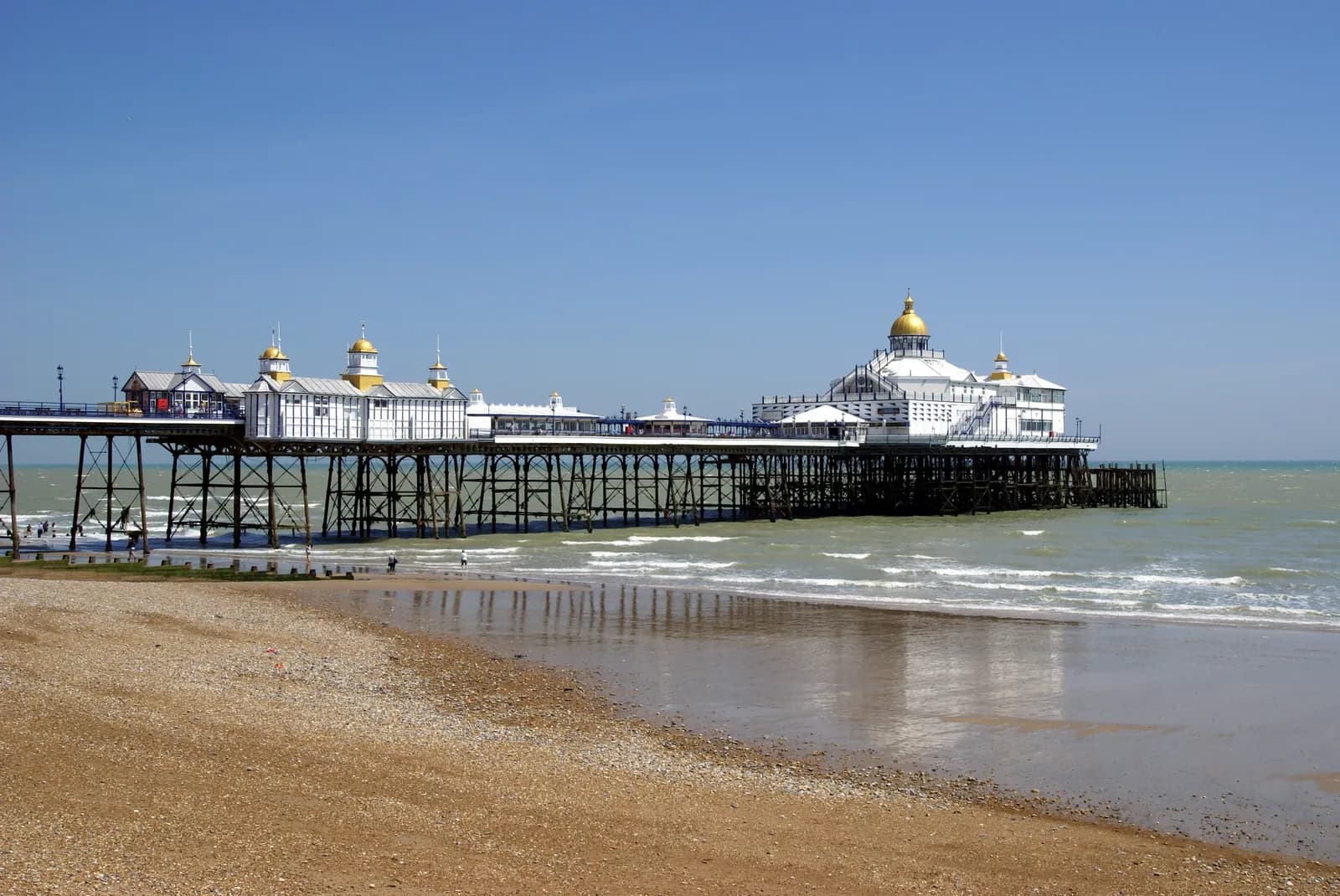 A long white pier with golden domes extends over the ocean from a sandy beach under a clear blue sky.