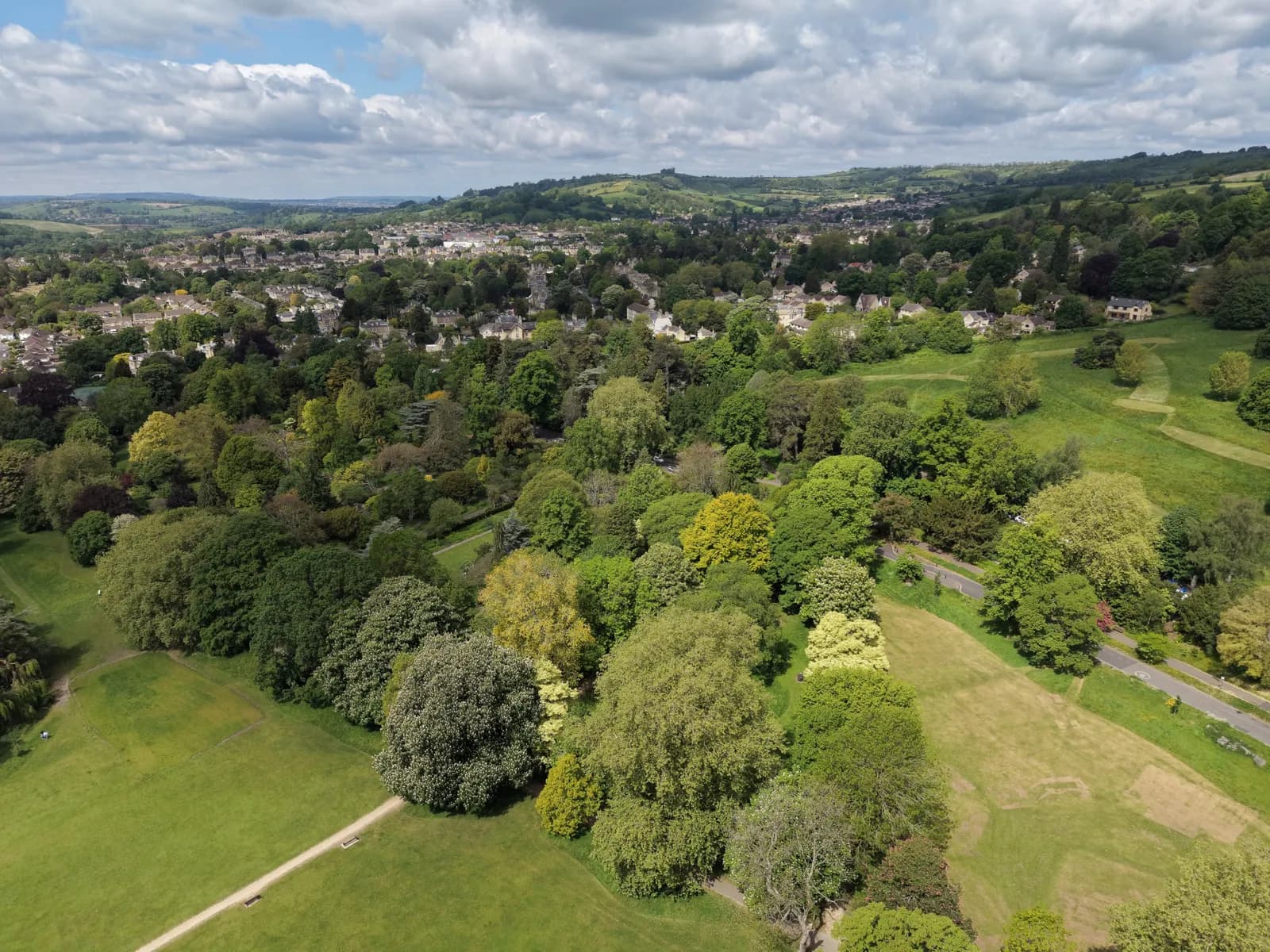 An aerial view of a verdant landscape with rolling green hills, dense clusters of trees, and a town nestled in the background under a cloudy sky.