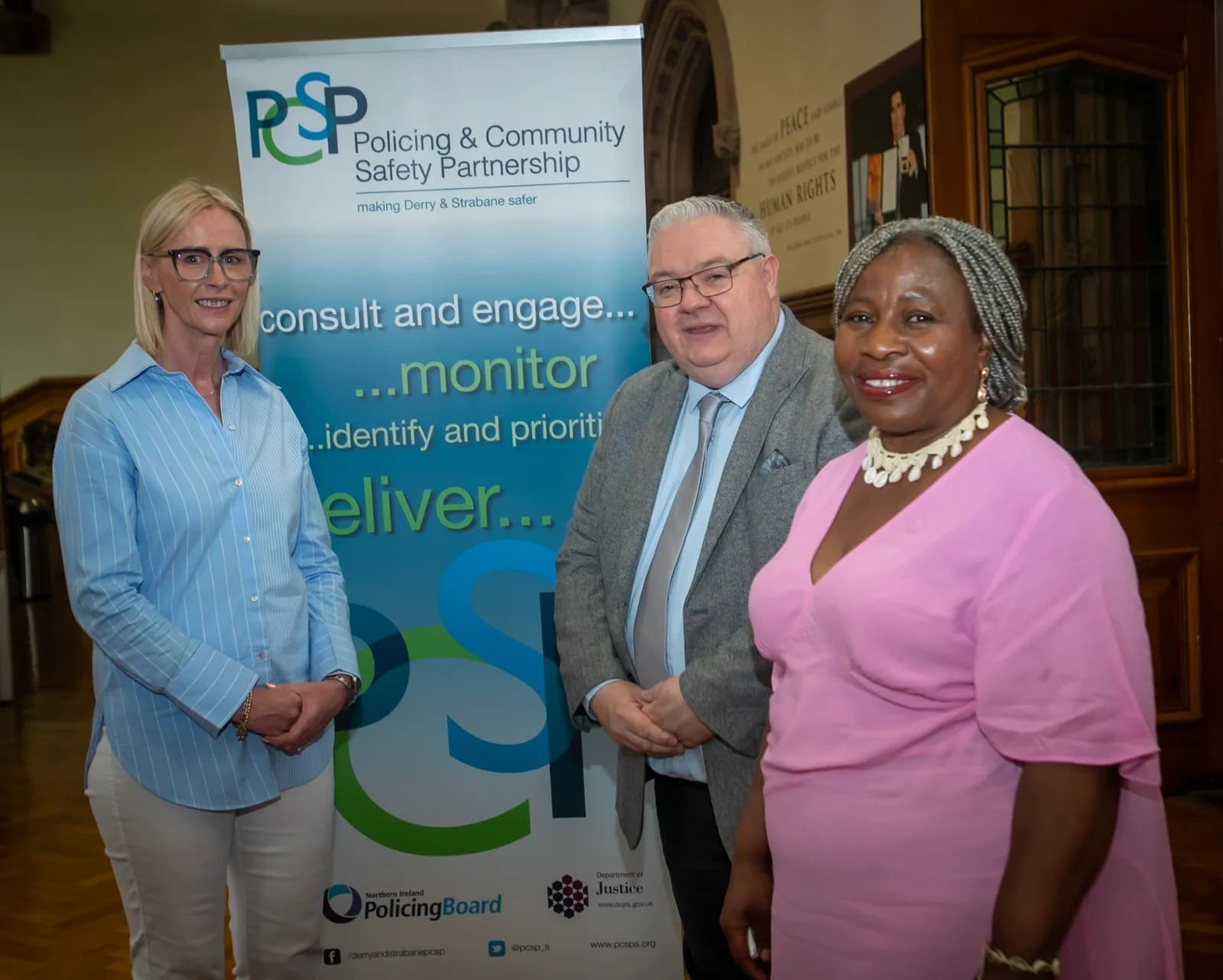 Three people stand in front of a banner for the Policing & Community Safety Partnership; a woman in a blue striped shirt is on the left, a man in a gray suit is in the middle, and a woman in a pink dress is on the right.