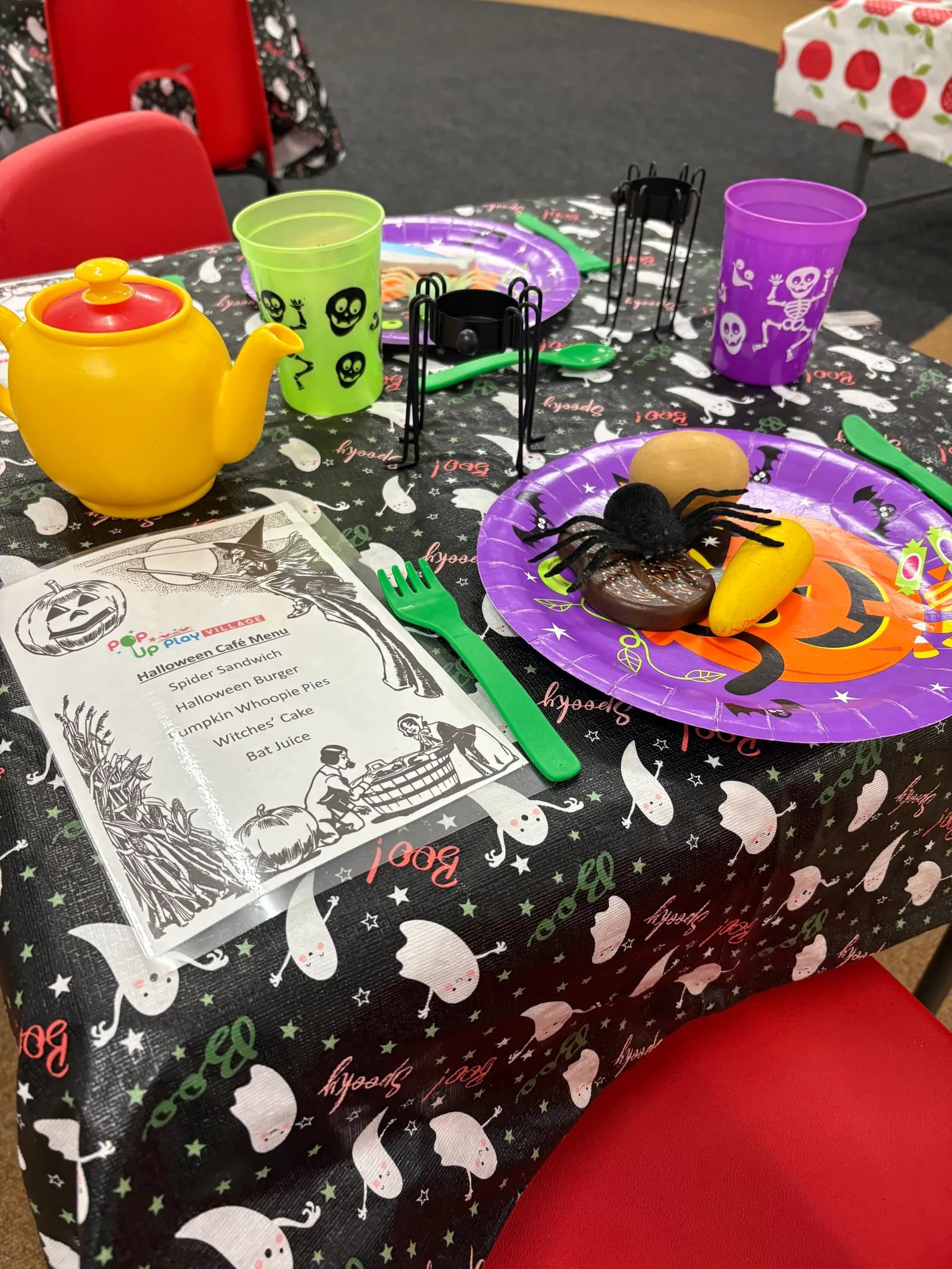 A table is set for a Halloween party with a spooky tablecloth, orange and purple plates, green utensils, spider decorations, and a yellow teapot.