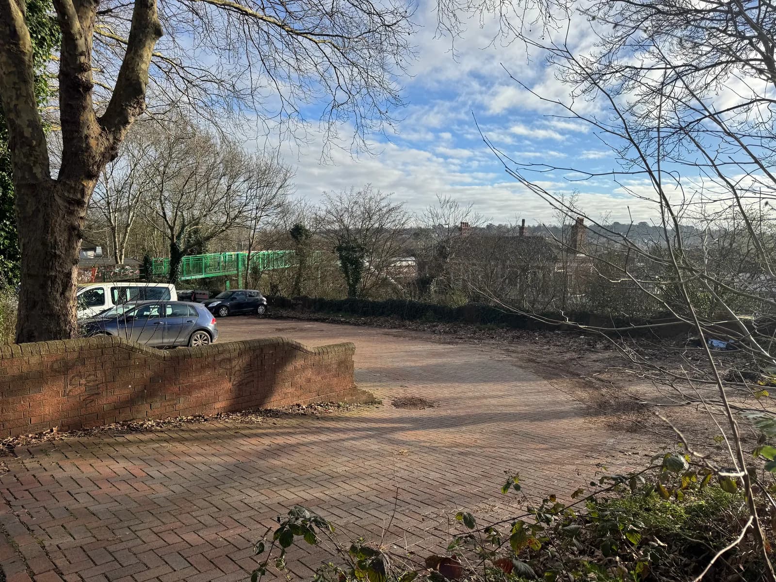 A paved area with a low brick wall on the left, cars and a green bridge in the background, and bare trees under a blue sky with clouds.