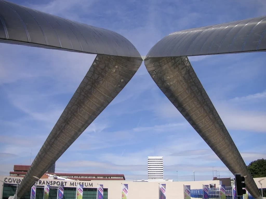 Two large, curved, metallic structures extend from the top corners, meeting in the center against a blue sky with scattered clouds, with buildings and a traffic light visible below.