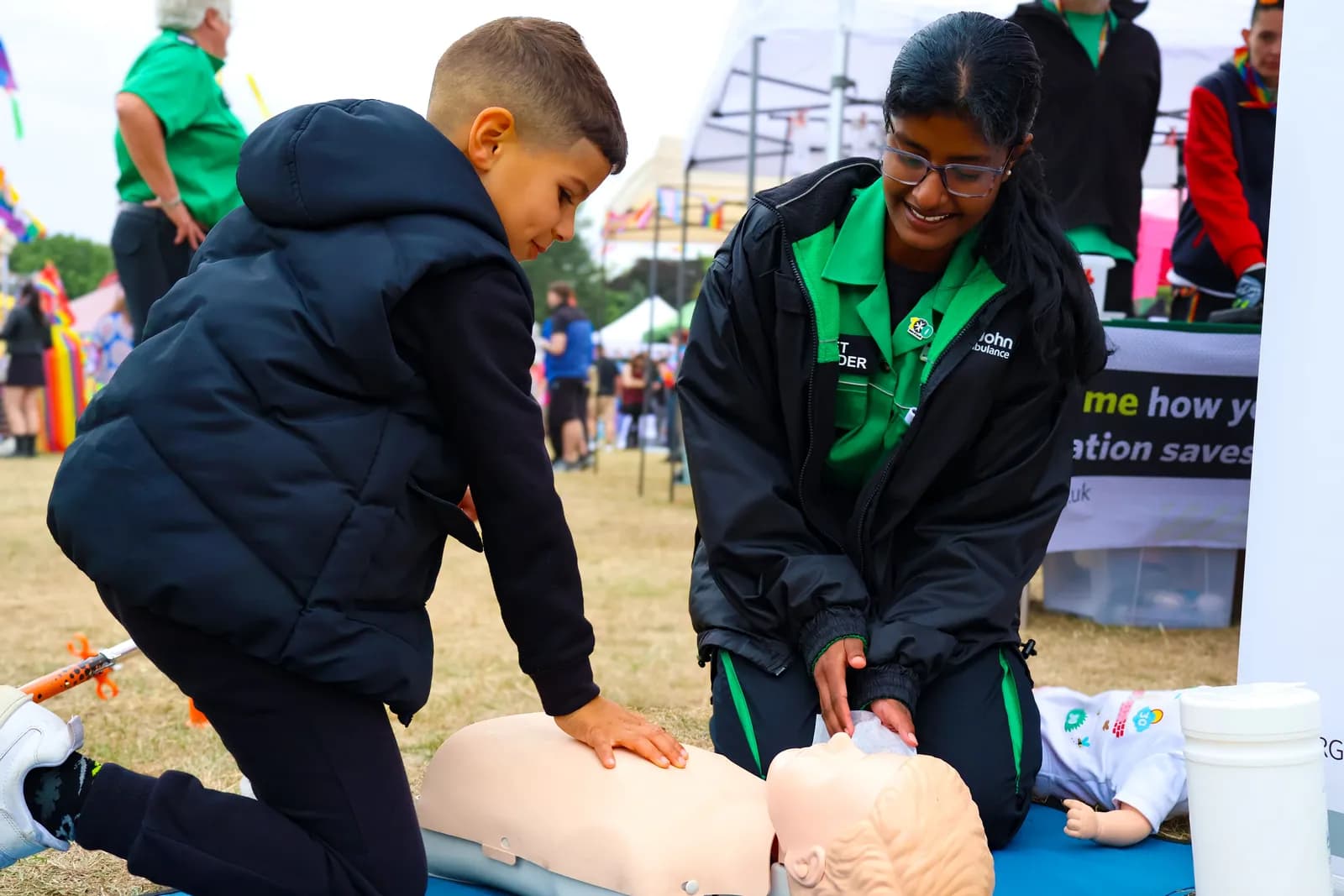A St. John Ambulance volunteer in a green and black uniform smiles while teaching a young boy how to perform CPR on a dummy.