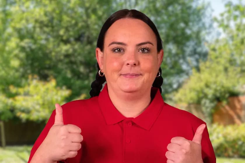 A woman with dark hair in pigtails, wearing a red polo shirt, smiles and gives two thumbs up against a blurred green background of trees and foliage.