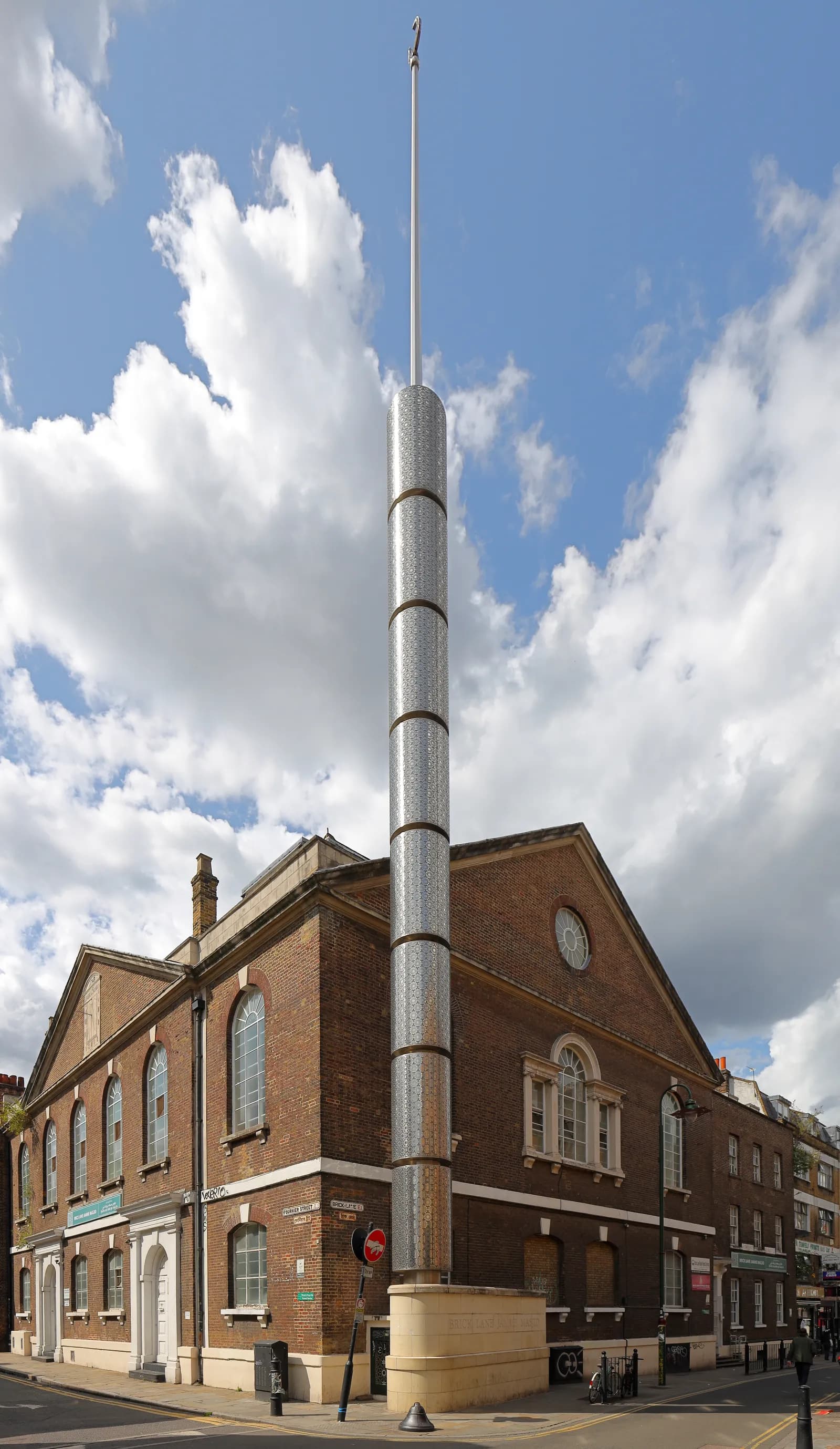 A tall, slender cylindrical metal column with a textured surface, extending upwards in front of a brick building with arched windows under a partly cloudy sky.