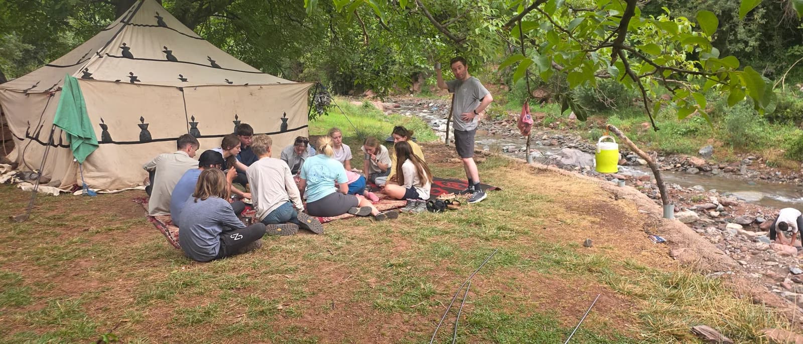 A group of young people are seated on the ground in a circle, some facing a man standing next to them, with a large tent to their left and a rocky stream to their right.