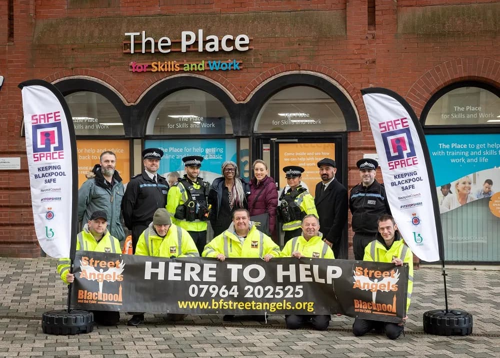 A group of people, some in police uniforms and others in high-visibility jackets, stand and kneel in front of a building called 'The Place for Skills and Work', holding a banner that reads 'HERE TO HELP'.