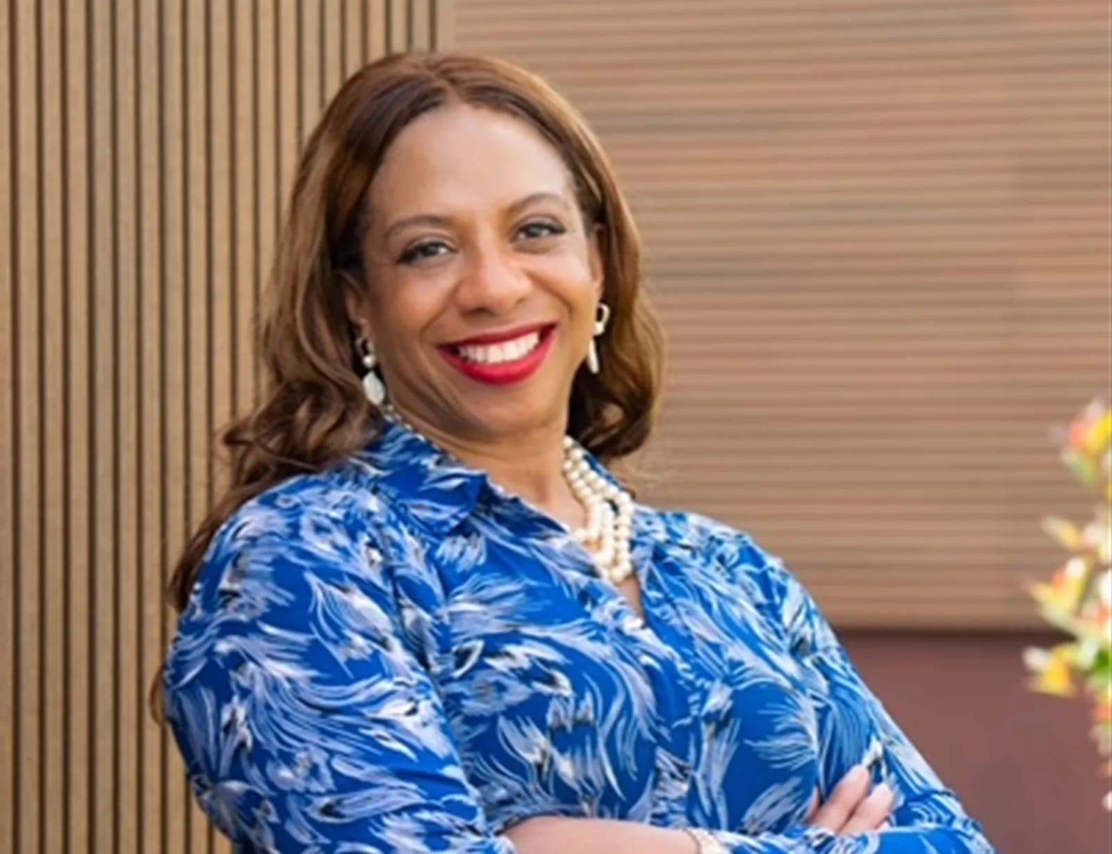 A woman with light brown hair, wearing a blue patterned shirt and pearl necklace, smiles against a background of wood paneling.