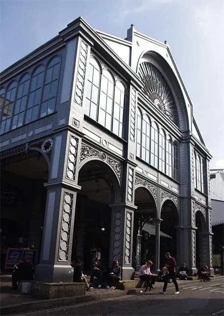 A grand, grey-painted building with Gothic-style arched windows, intricate architectural details, and several archways on the ground floor with people sitting underneath.