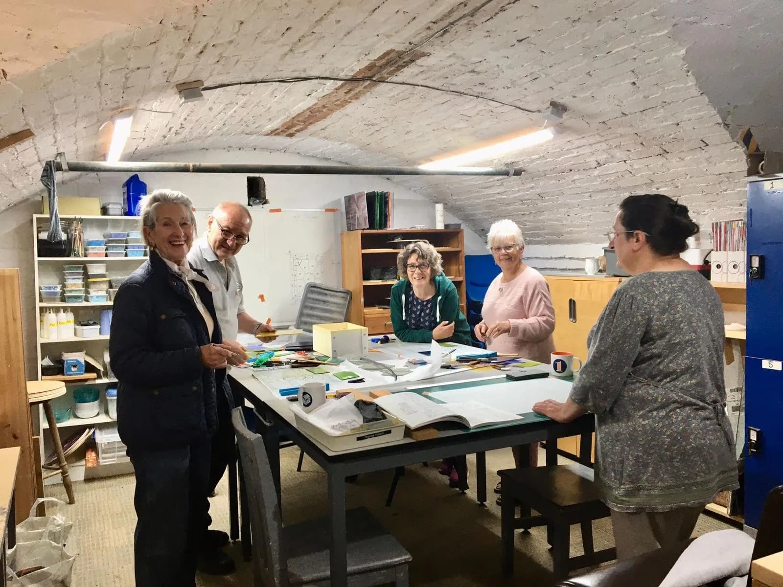 A group of five people of various ages are gathered around a table with craft supplies in what appears to be a workshop or studio with arched brick ceilings.