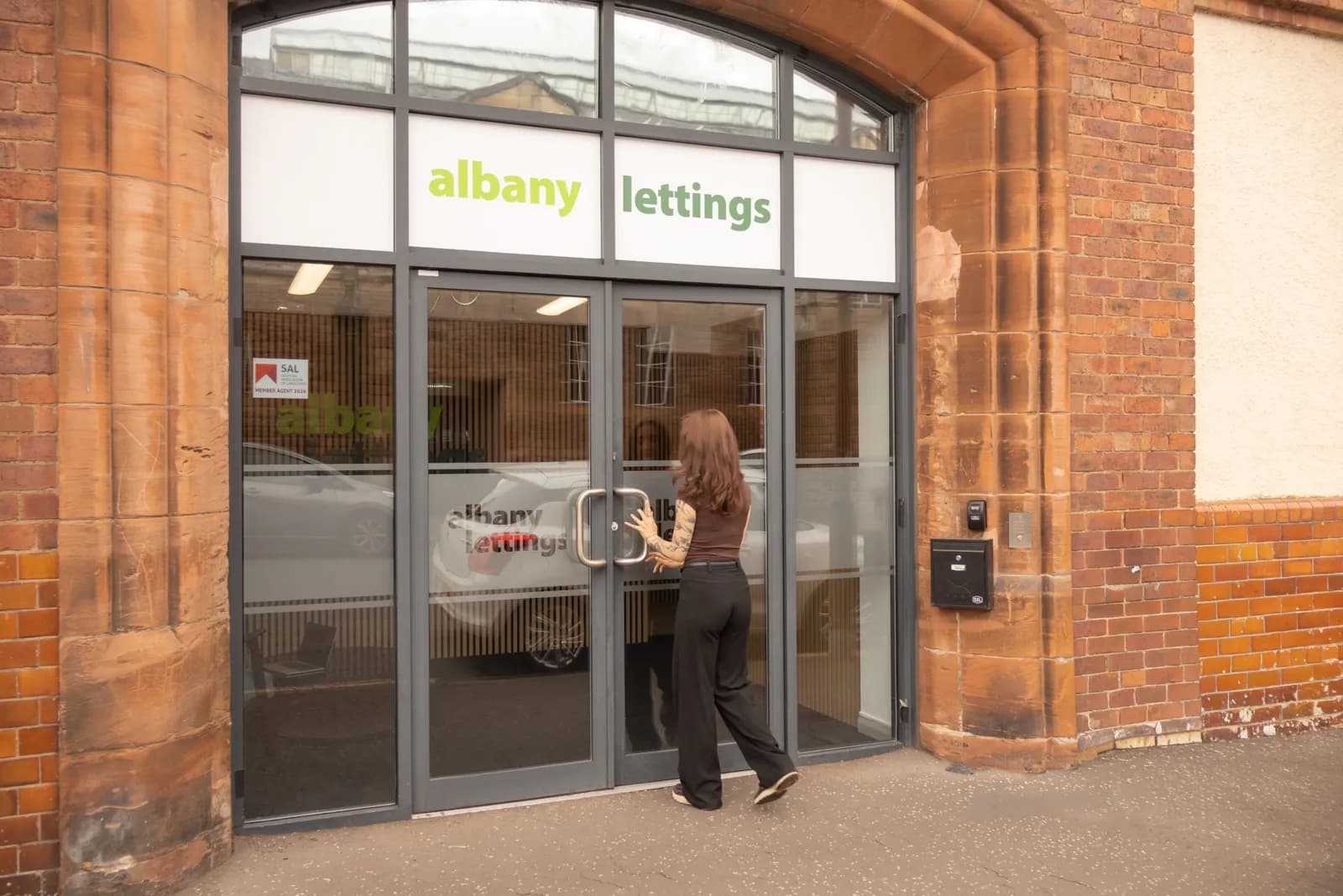 A person with long brown hair, wearing a brown top and black pants, is about to open the glass doors of an Albany Lettings building.