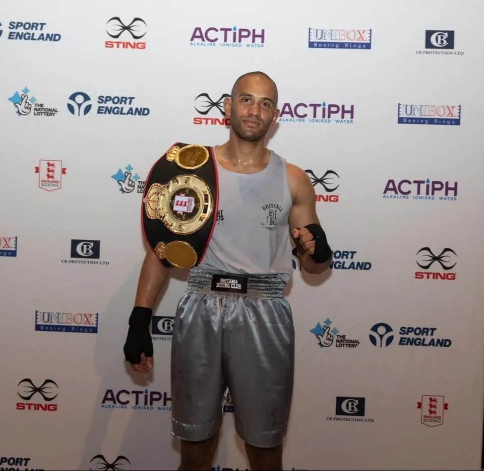 A male boxer with a shaved head and a light gray tank top holds a large gold championship belt over his shoulder, standing in front of a white wall with various sponsor logos.