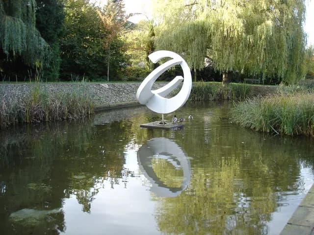 A modern white abstract sculpture with a circular form stands in the middle of a calm pond, surrounded by lush green foliage and trees, with its reflection visible in the water.