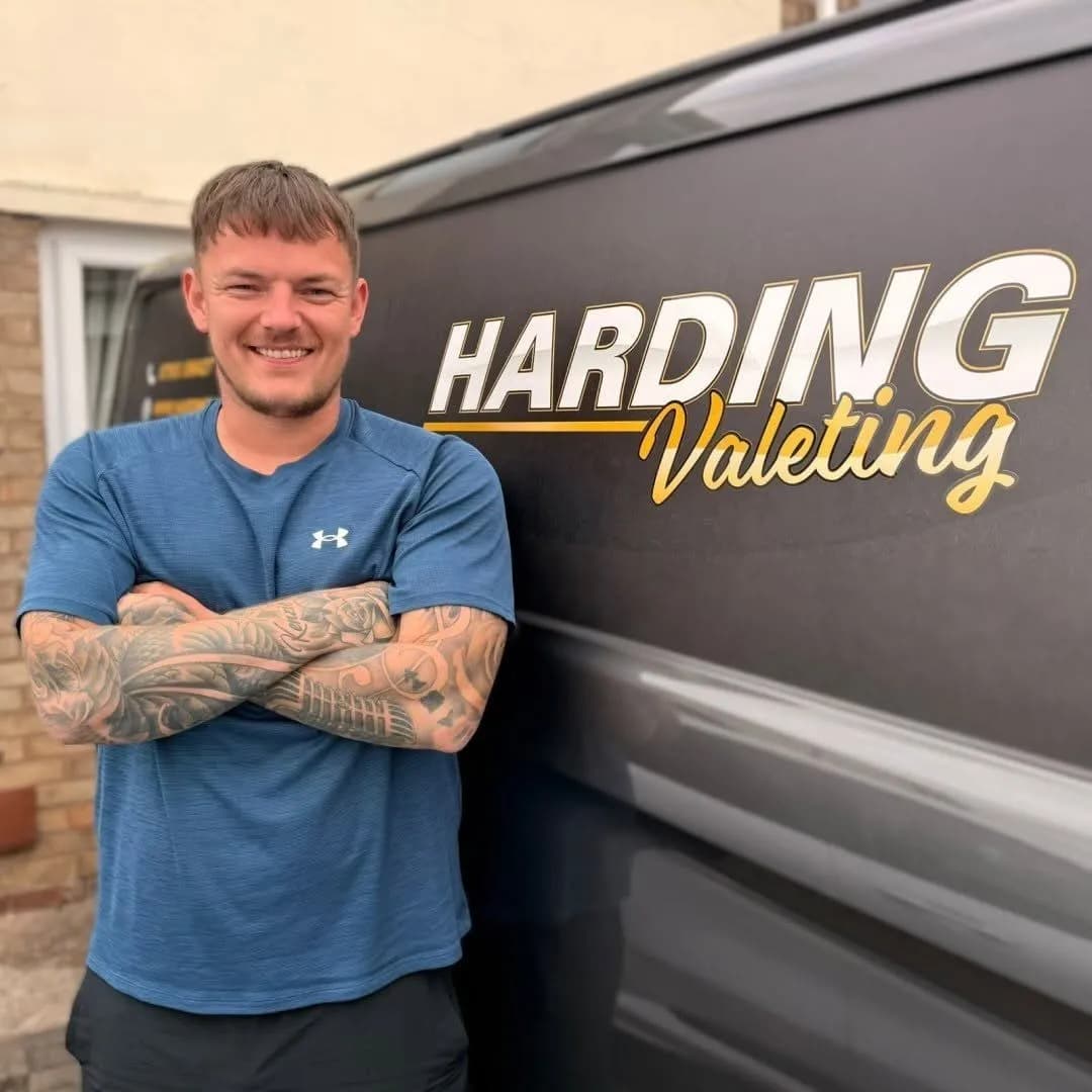 A man with short brown hair, wearing a blue shirt, stands smiling with his arms crossed in front of a dark van with 'HARDING Valeting' written on its side.