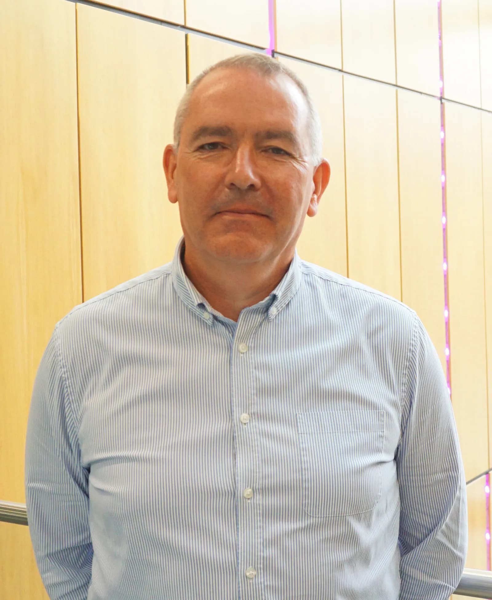 A man with short grey hair and a light blue striped button-up shirt standing in front of a light brown paneled wall.