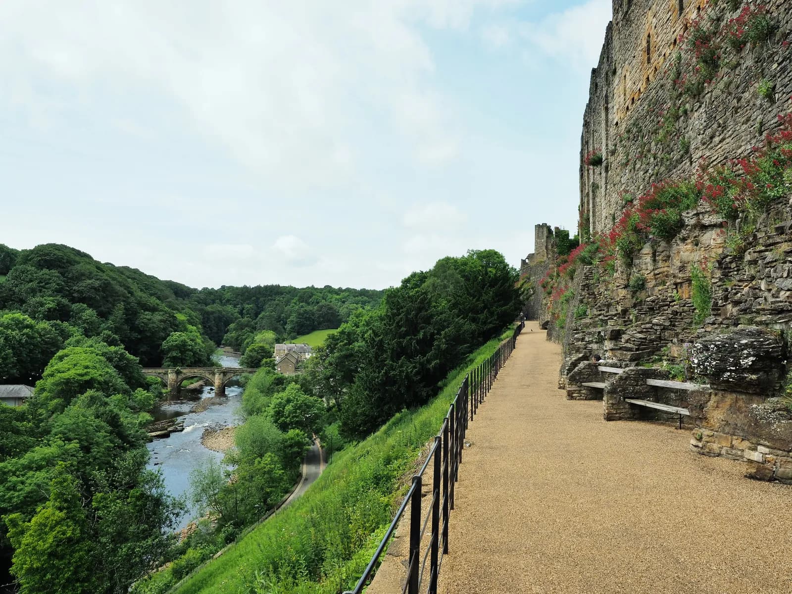 A long, gravel pathway with a black metal railing on the left and a stone castle wall on the right, overlooking a green valley with a river and an old stone bridge.