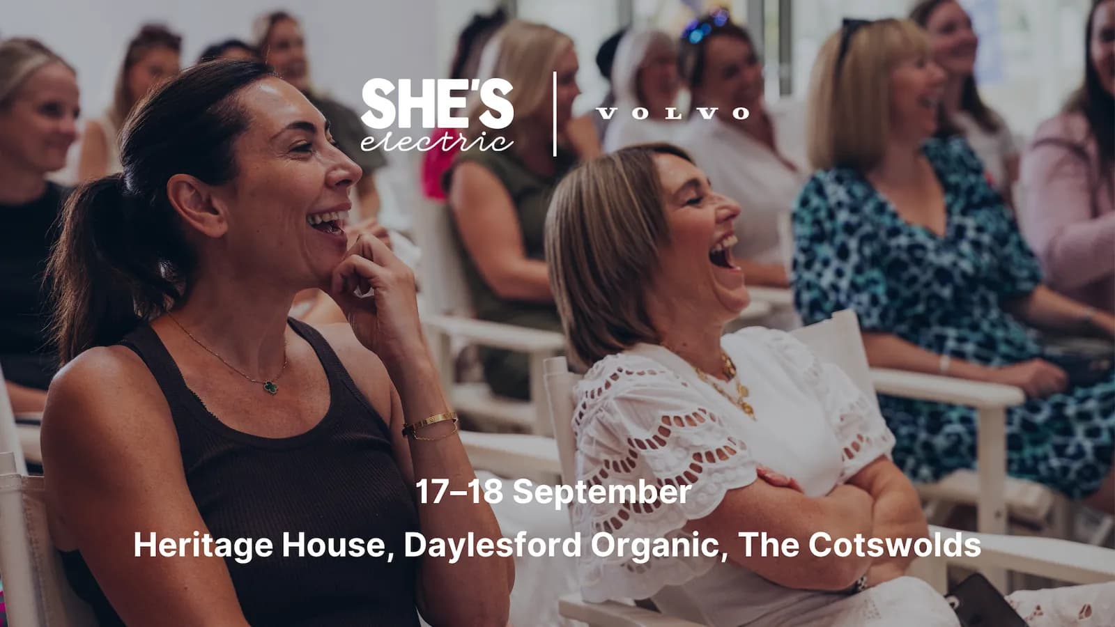 Two women laugh heartily while seated in an audience, with event details overlayed.