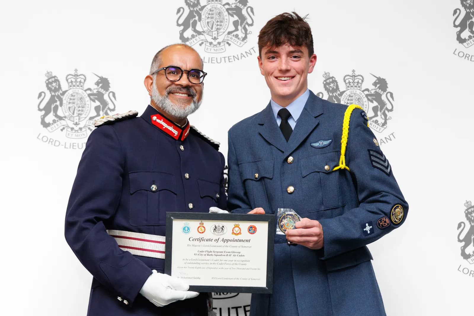 Two men, one in a dark uniform and the other in a light blue uniform, both smiling, holding a framed certificate and a medal.