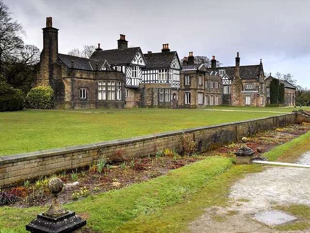 A long, historic stone and half-timbered manor house with multiple chimneys and gabled roofs overlooks a manicured green lawn and a flowerbed separated by a low stone wall.