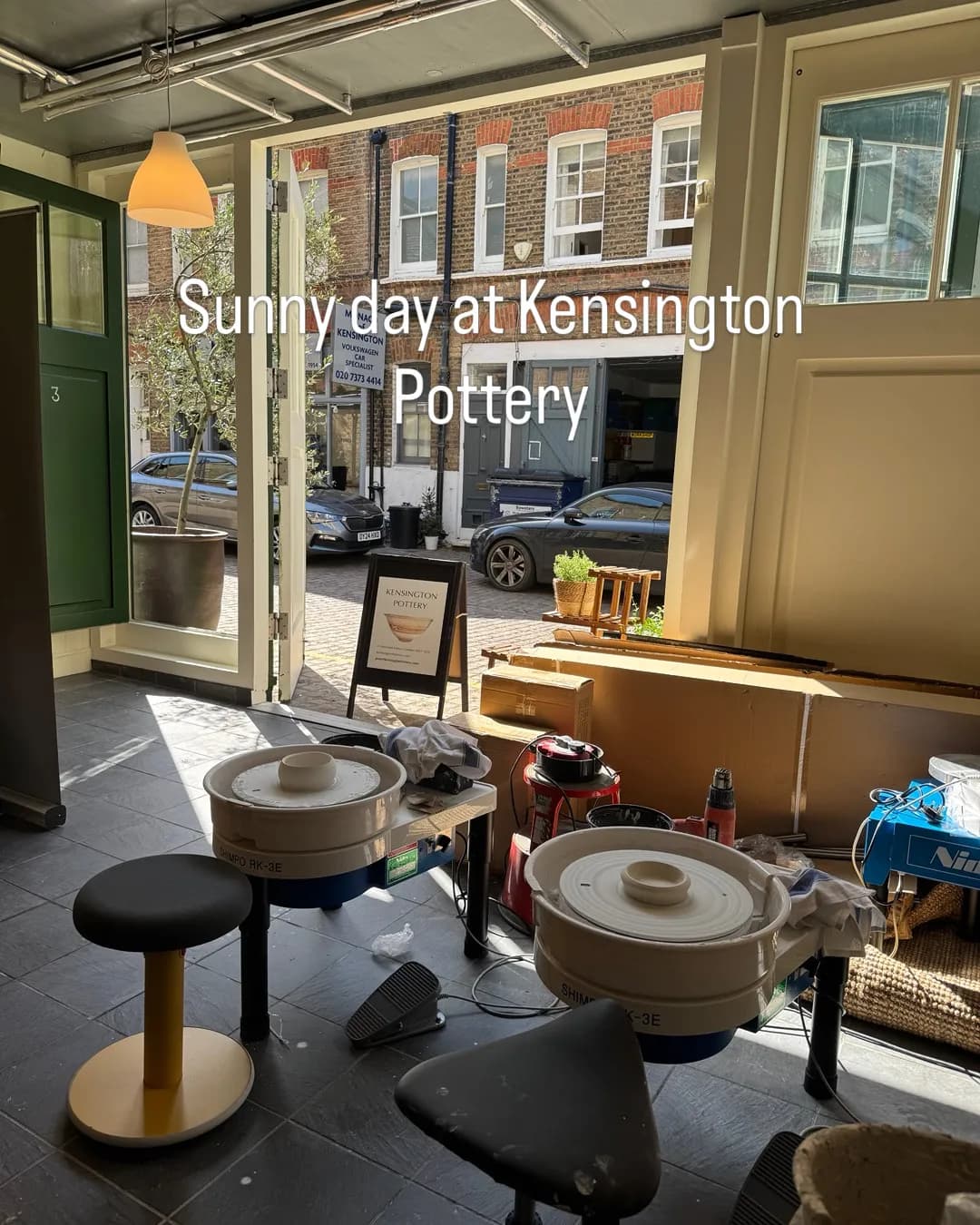 A brightly lit pottery studio with two pottery wheels and stools, looking out through an open doorway to a street with brick buildings and parked cars.