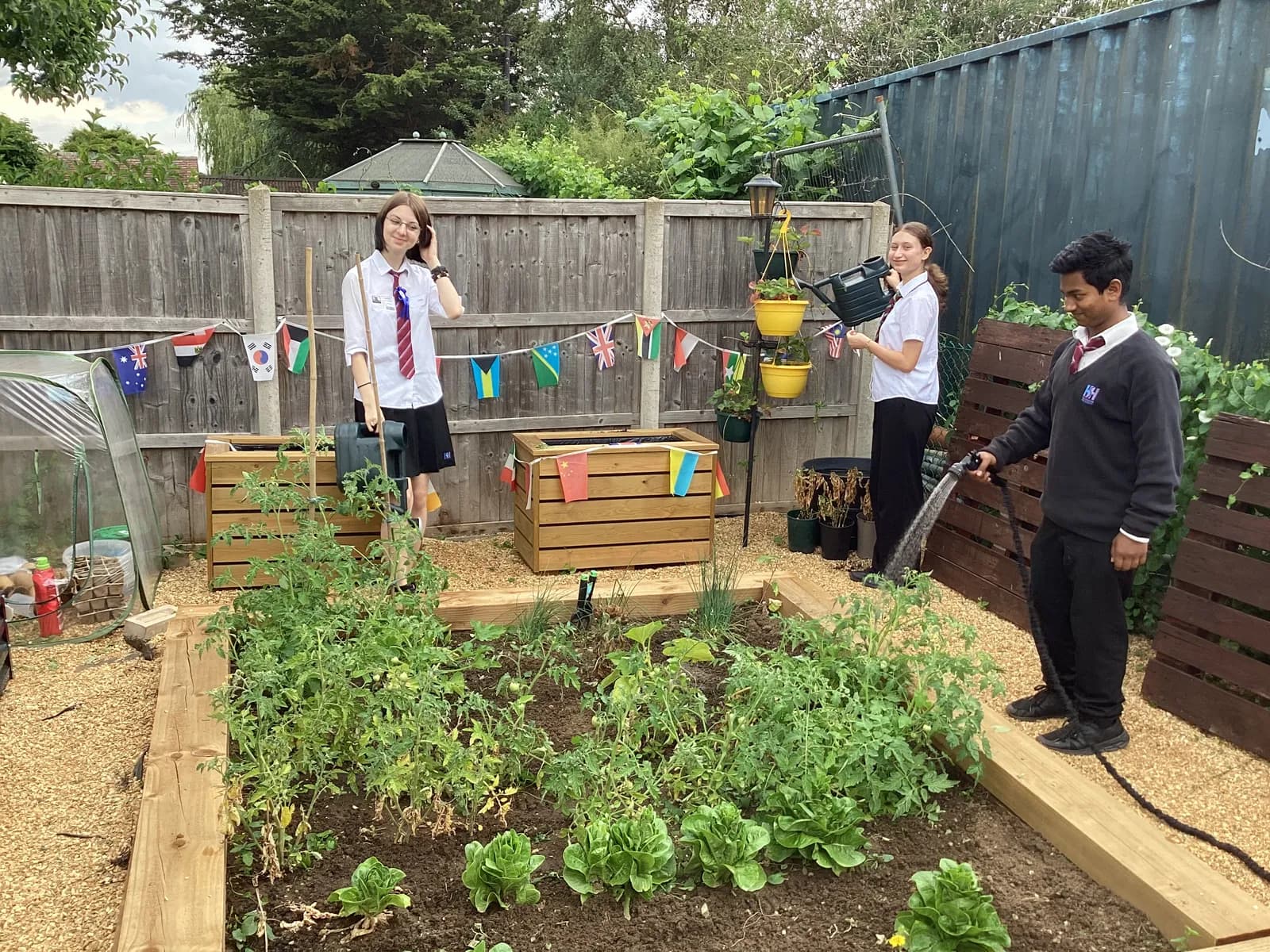 Three students in school uniform are tending to a garden, with one girl standing near a planter, another girl watering hanging plants, and a boy watering plants in a raised bed.
