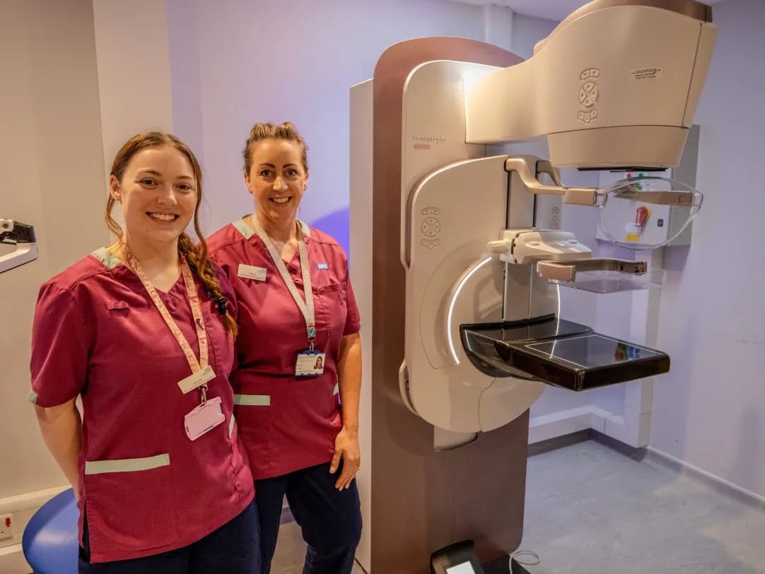 Two women wearing maroon scrubs with light green trim stand smiling next to a large mammography machine in a medical room.