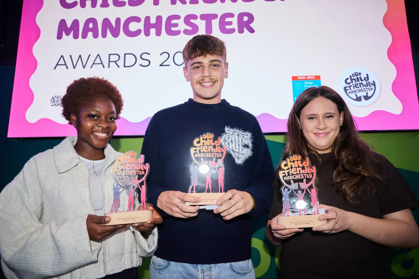 Three young adults, two women and one man, stand smiling and holding "Our Child Friendly Manchester" awards, with a large sign in the background.