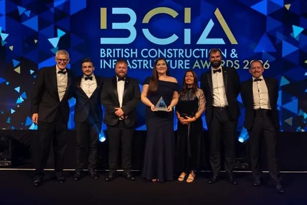 A group of seven people, six men and one woman, all dressed in formal wear, stand on a stage in front of a blue digital screen displaying the 'BCIA British Construction & Infrastructure Awards' logo, with the woman in the center holding a clear pyramid-shaped award.