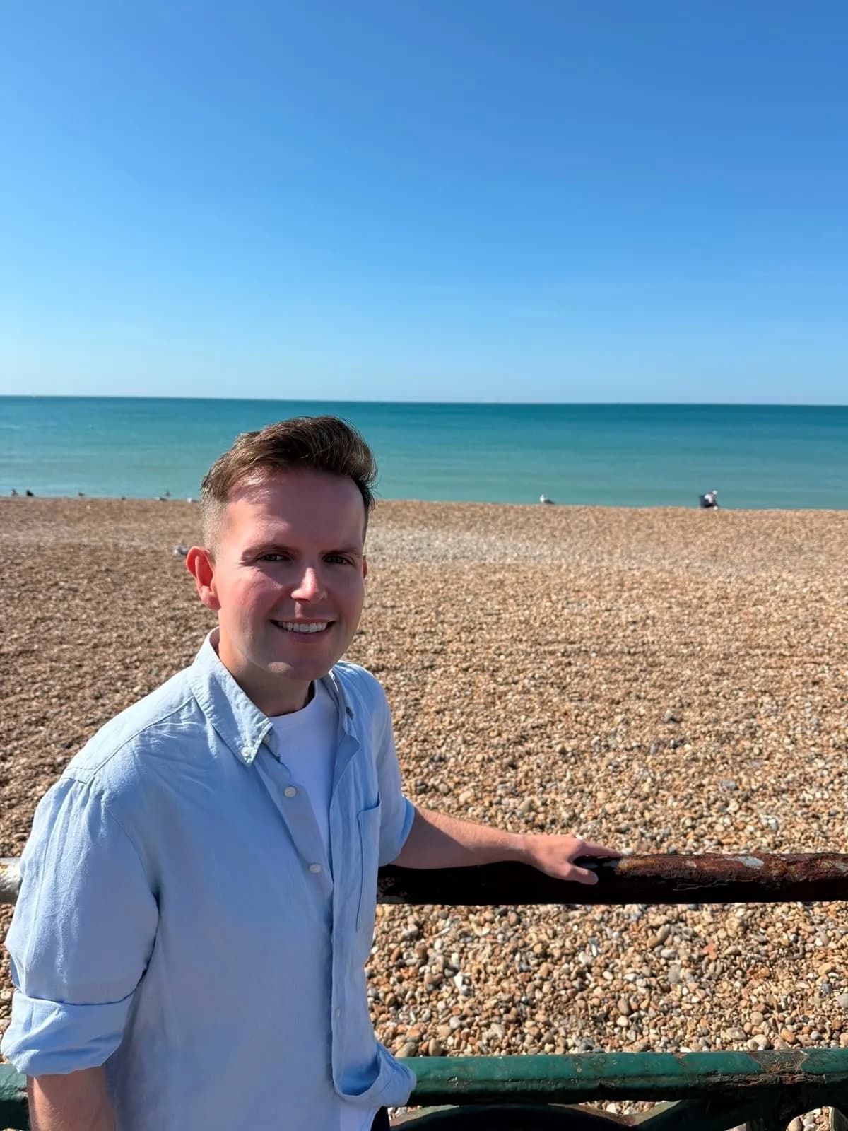 A man with short brown hair and a light blue shirt smiles at the camera while standing on a pebble beach with the ocean and a clear blue sky in the background.