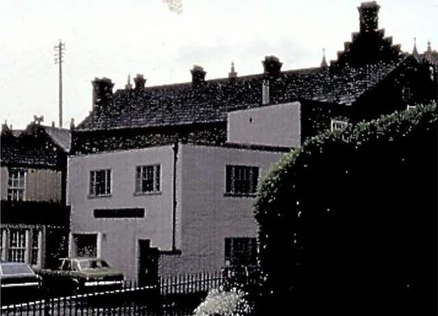A light-colored building with dark-tiled roofs, surrounded by trees and a few cars parked in front.