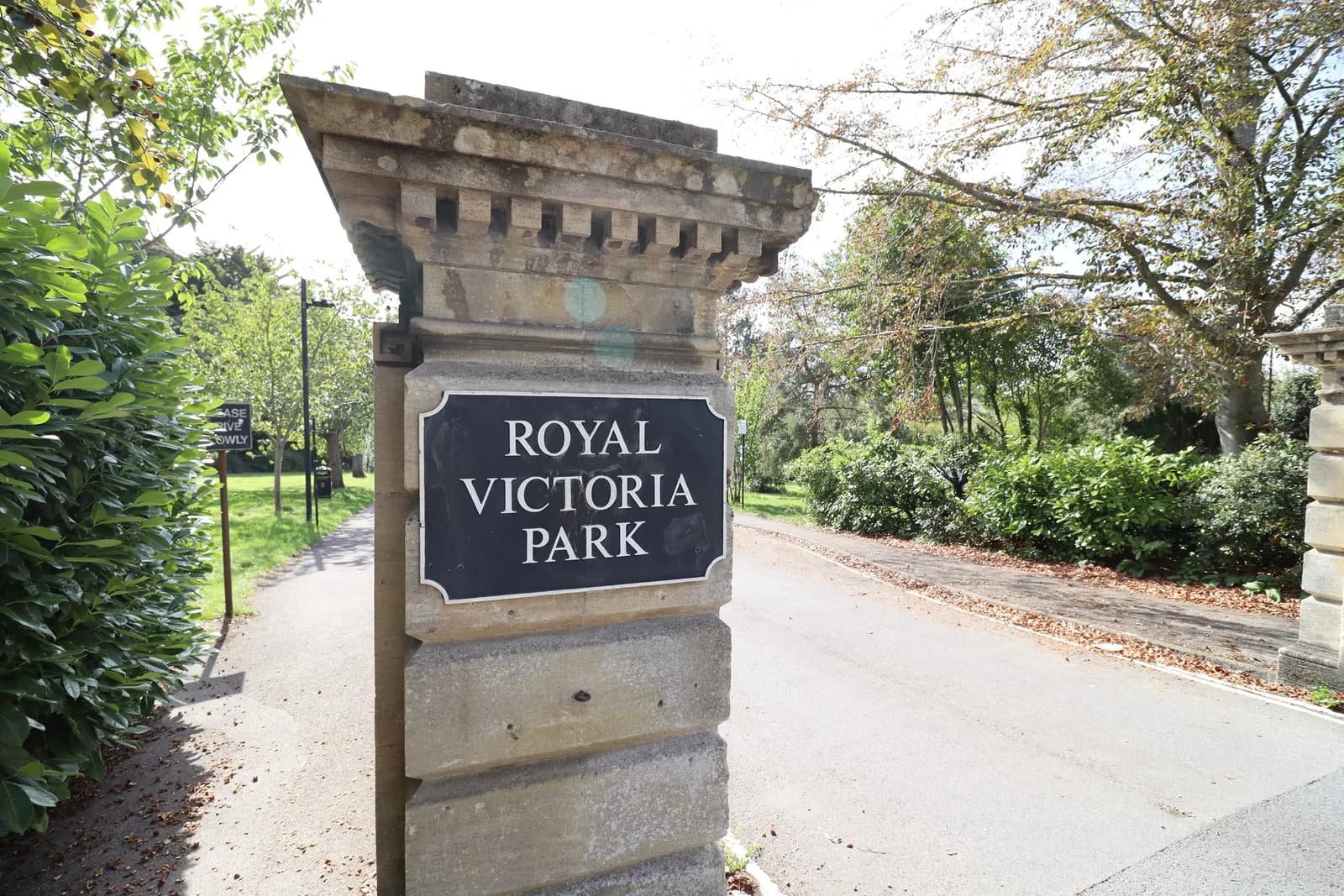 A large stone pillar with a black sign that reads "ROYAL VICTORIA PARK" in white letters stands beside a paved path and road, with green trees and bushes in the background under a bright sky.