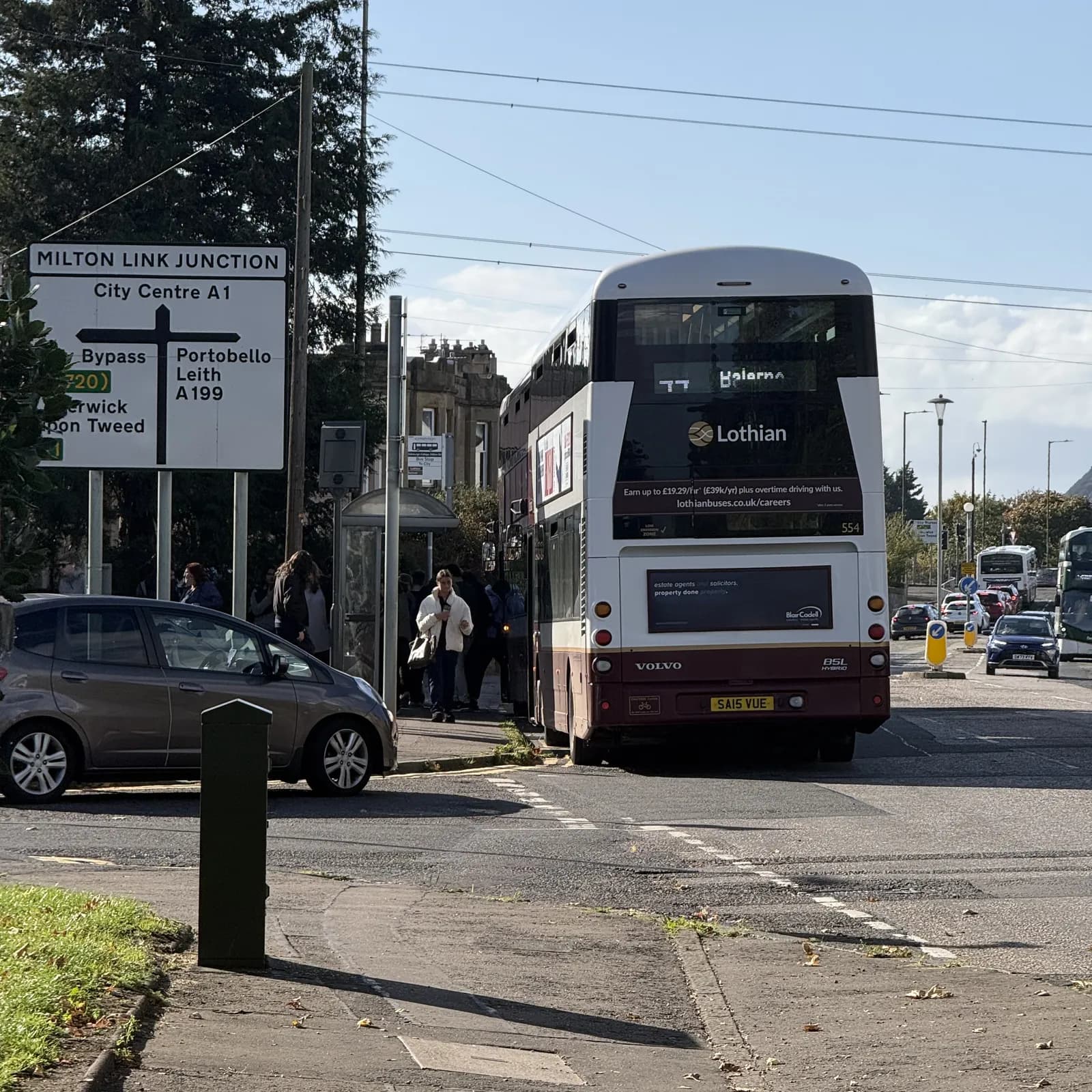 The rear of a white and maroon Lothian bus with "Balerno" displayed, is stopped at a bus stop on a sunny day with cars and a road sign nearby.