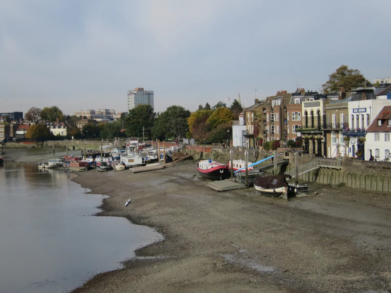 Several small boats are moored on a muddy riverbank next to a row of historical buildings, with a distant cityscape visible under an overcast sky.