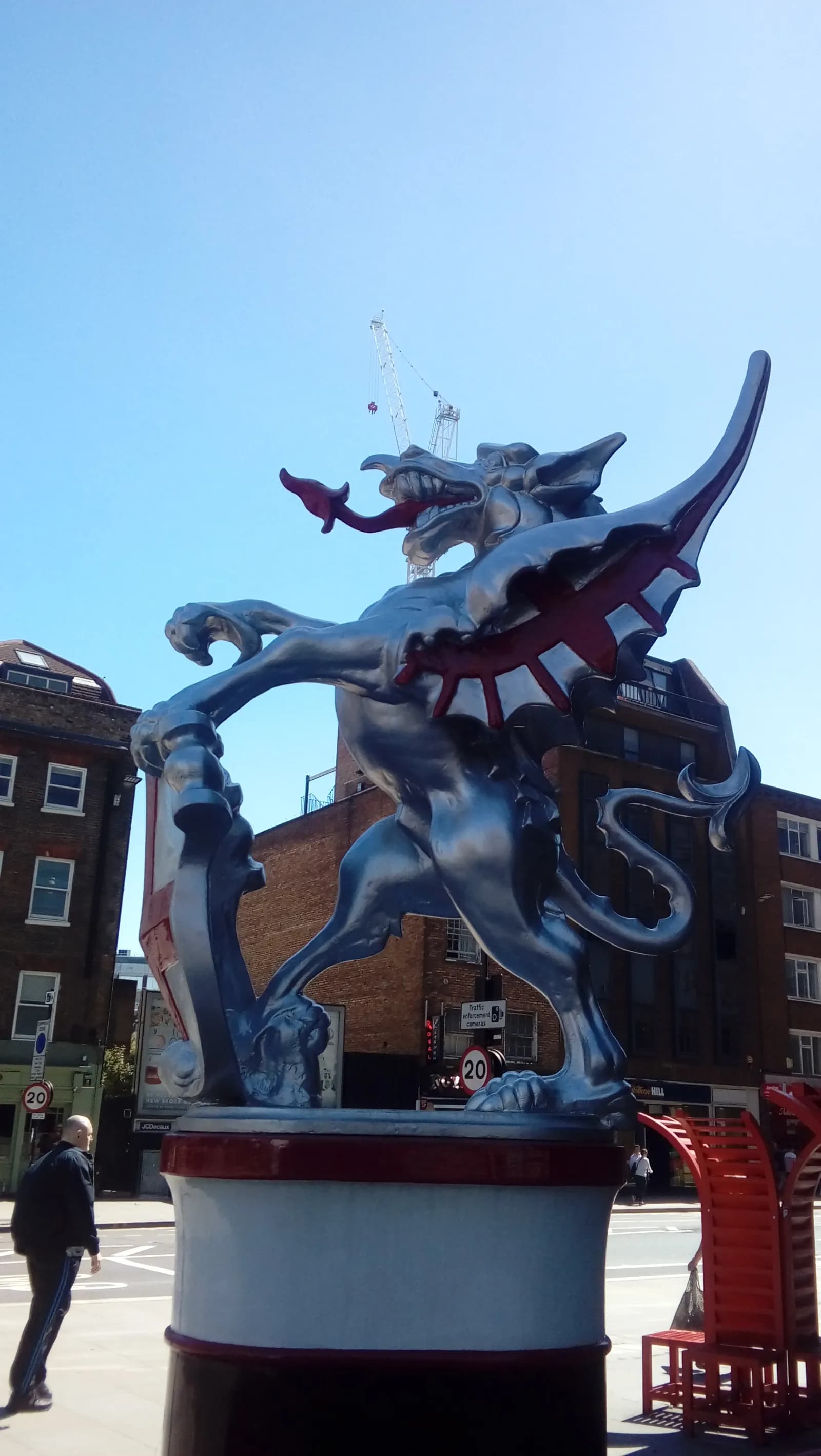 A large silver griffin statue with red accents stands prominently on a pedestal against a clear blue sky, with buildings and a construction crane in the background.