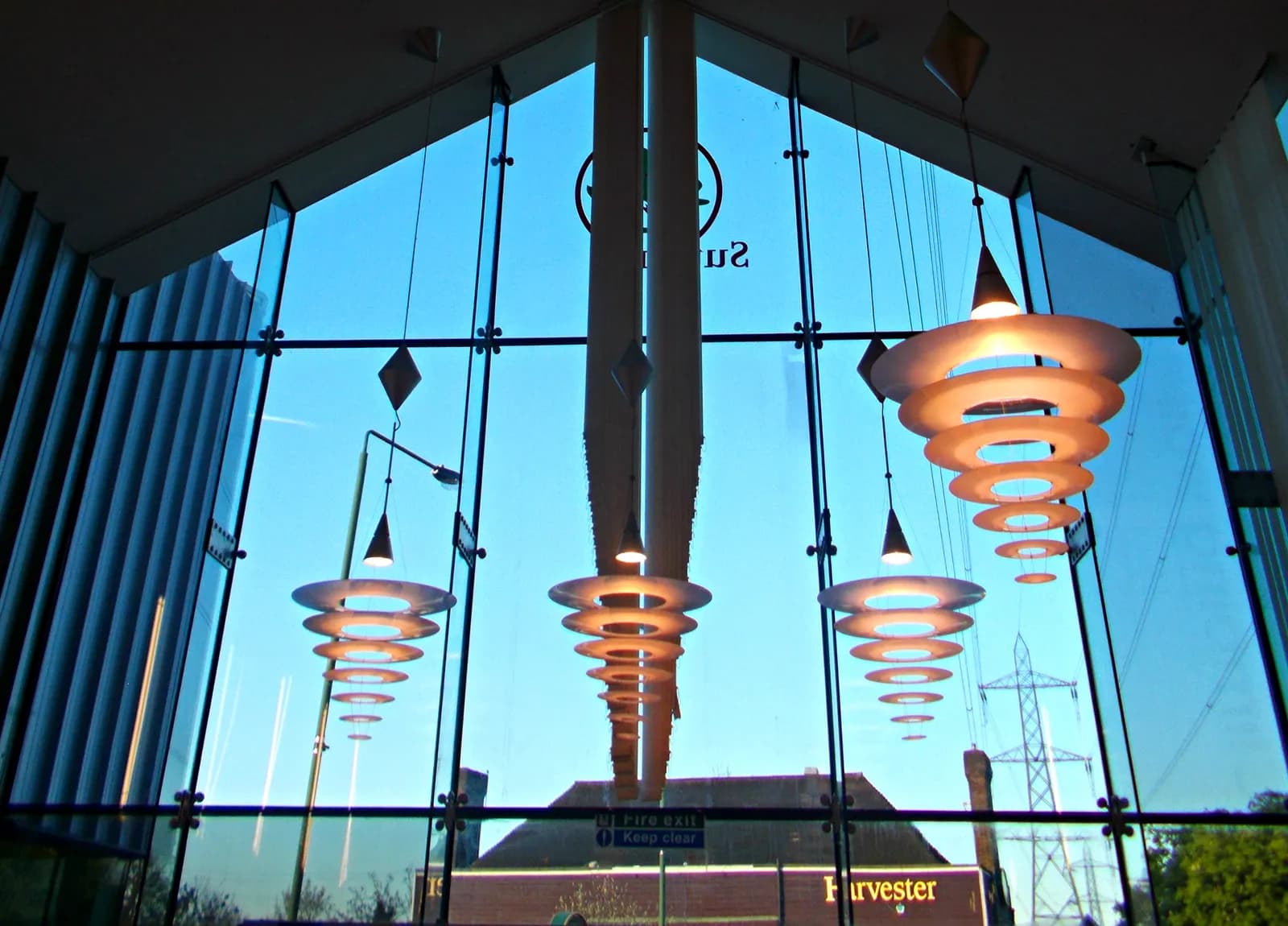 A view through large glass windows of a building, showing three groups of circular, tiered lights hanging from the ceiling, with a bright blue sky and distant buildings and a power line tower in the background.