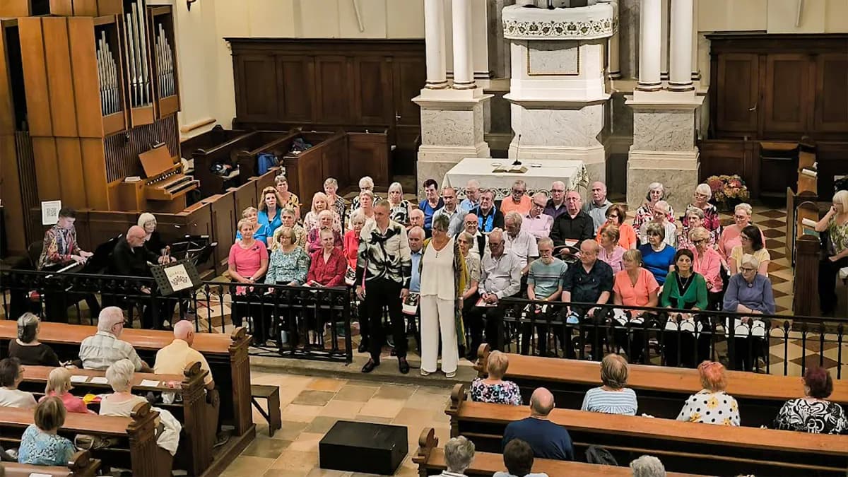 A choir stands and sits in a church, with a pianist and cellist performing to the left, and an audience visible in the pews in the foreground.