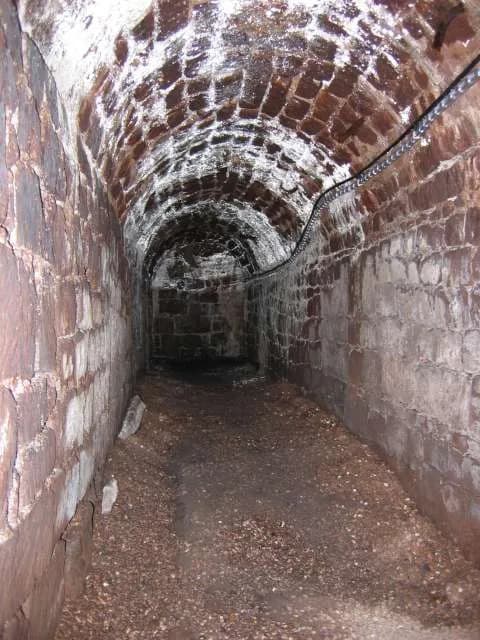 A long, dark underground tunnel with a vaulted brick ceiling and walls, and a loose gravel floor.
