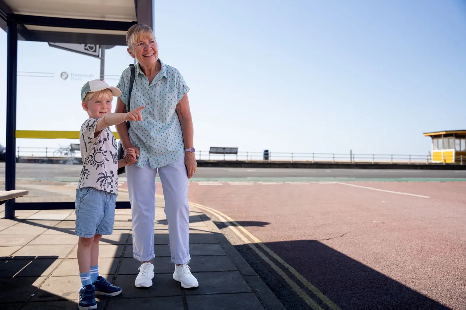 A happy grandmother and her young grandson holding hands, standing at a bus stop with the grandson pointing at something in the distance.