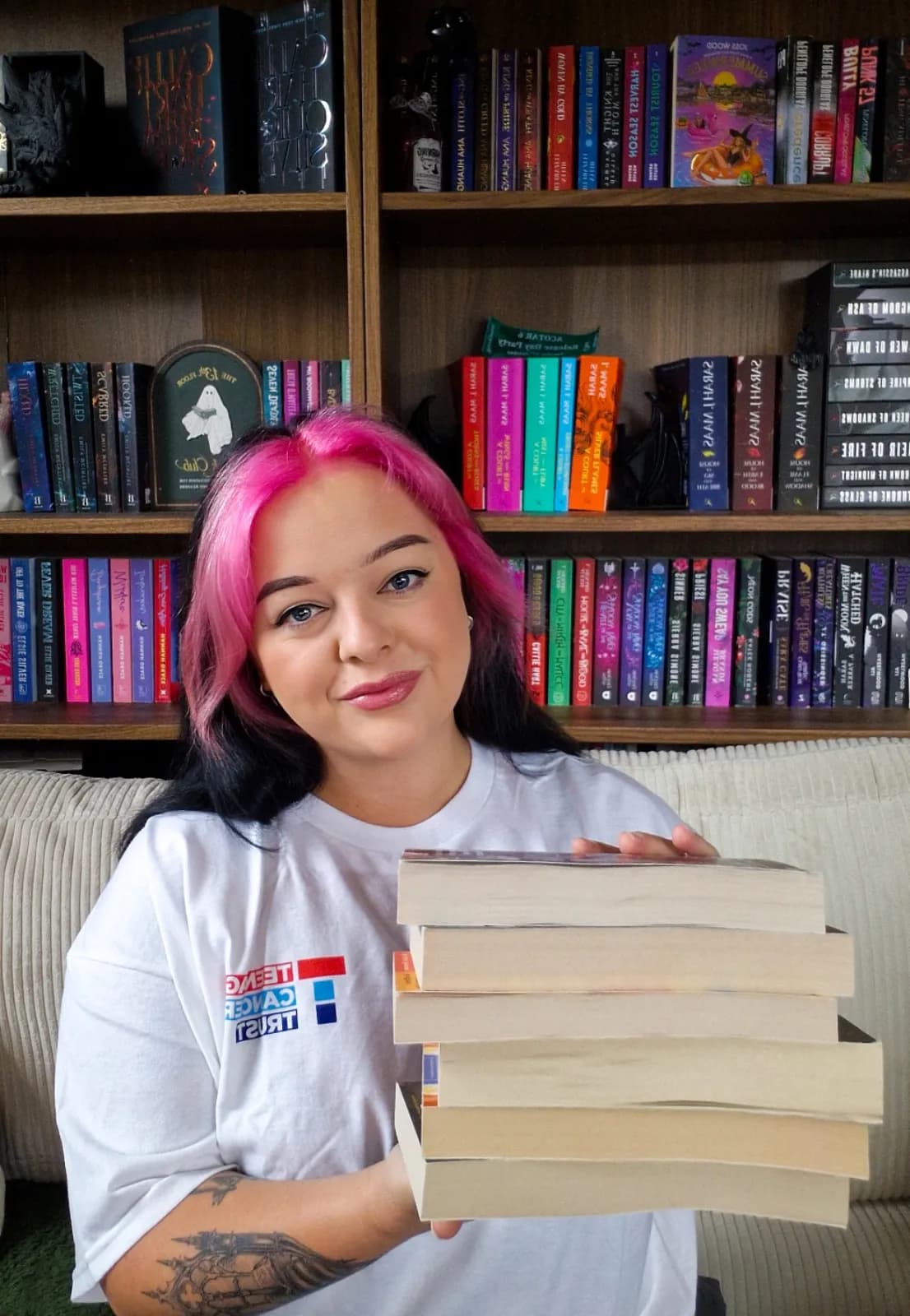 A woman with bright pink hair holds a stack of books, smiling at the viewer in front of a bookshelf filled with various titles.