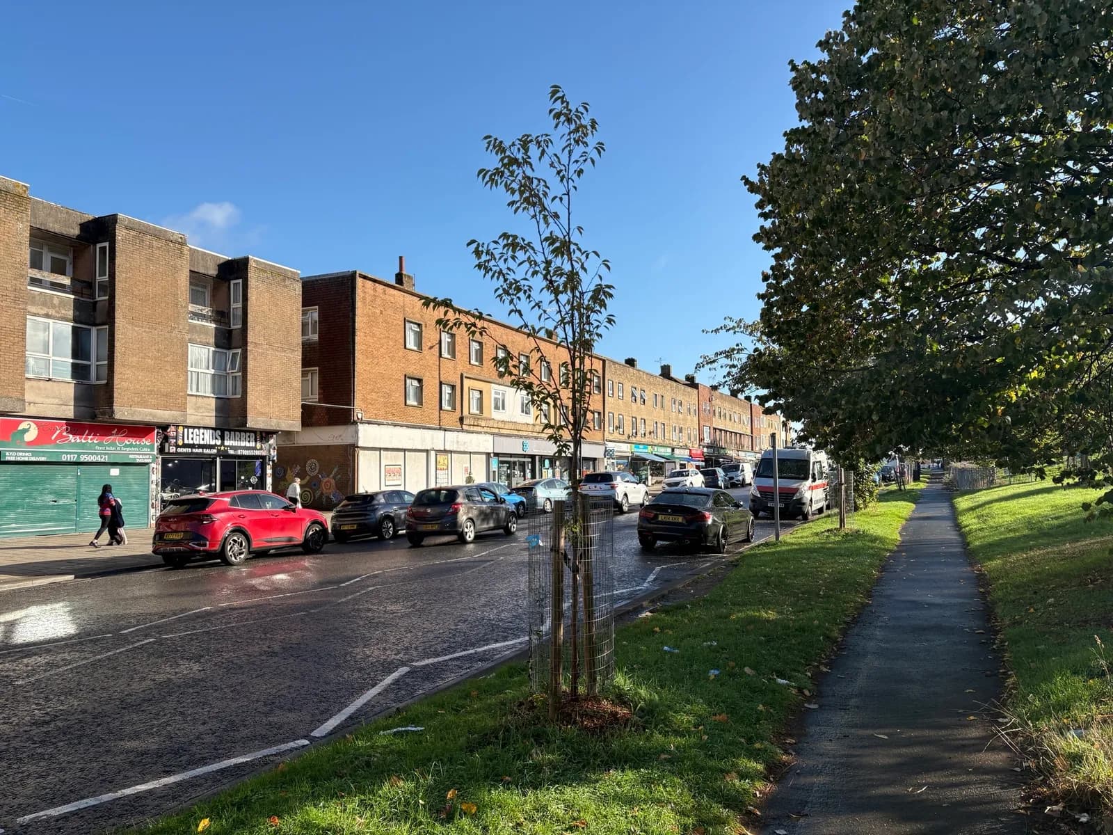 A street view with brick buildings housing shops on one side and a grassy area with trees and a sidewalk on the other, with cars parked along the road and a person walking.