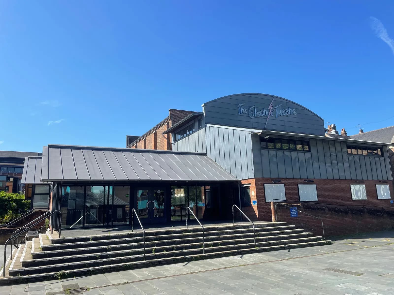 The exterior of The Electric Theatre building under a clear blue sky, featuring brick walls, grey metallic cladding, and a prominent entrance with steps leading up to glass doors.