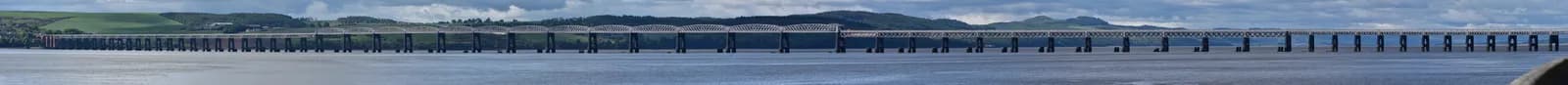A long railway bridge stretches across a wide body of water, with rolling green hills and mountains in the background under a cloudy sky.