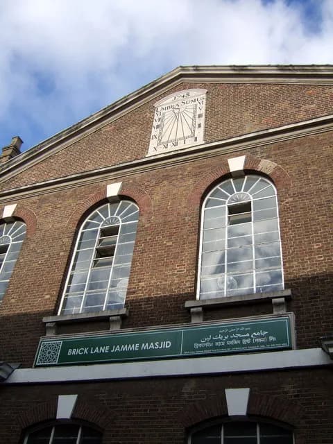 A close-up view of the Brick Lane Jamme Masjid with its distinctive architecture, including a sundial on the pediment and large arched windows, under a partly cloudy sky.