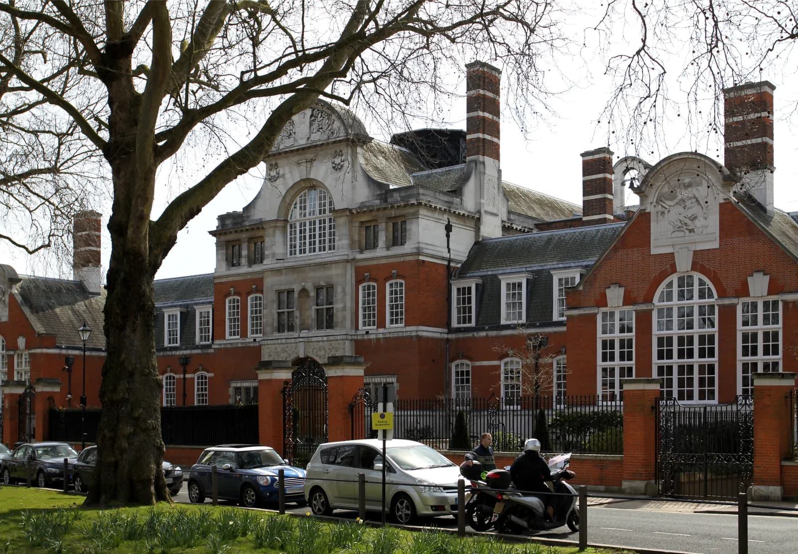 A grand, red brick building with white architectural details, tall windows, and multiple chimneys, viewed from across a road with parked cars and a large bare tree.