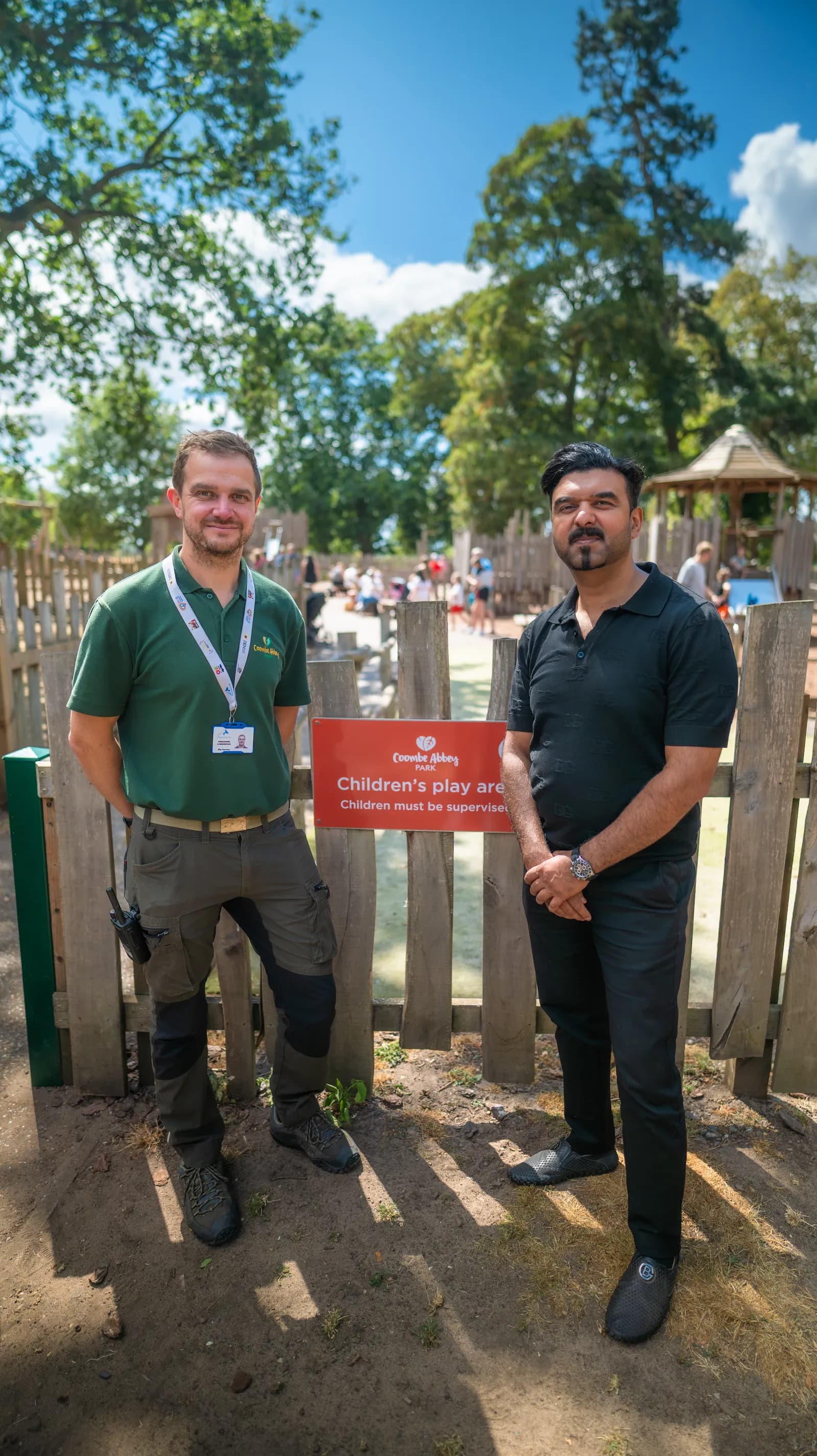 Two men, one in a green polo and cargo pants and the other in a black polo and black pants, stand on a dirt path in front of a wooden fence with a red sign, in an outdoor play area under a blue sky.