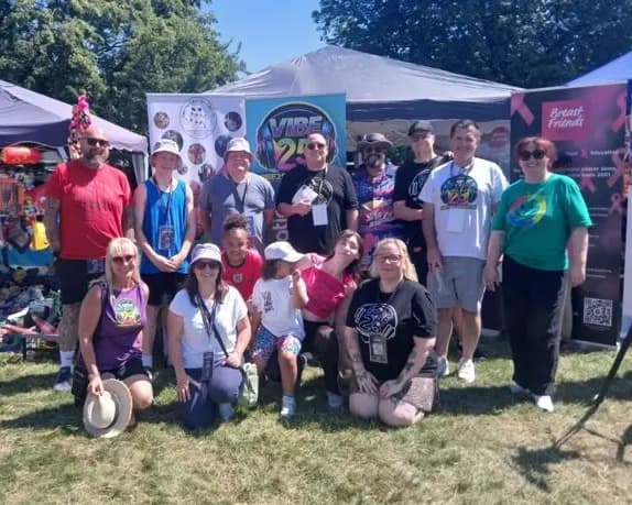 A diverse group of people of all ages stand and kneel in front of tents and banners on a sunny day.