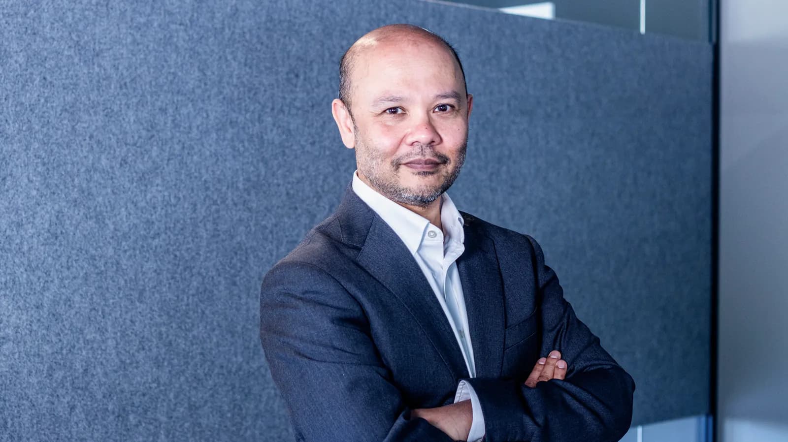 A man with a bald head and a short beard wears a dark suit and white shirt, looking directly at the viewer with a slight smile.