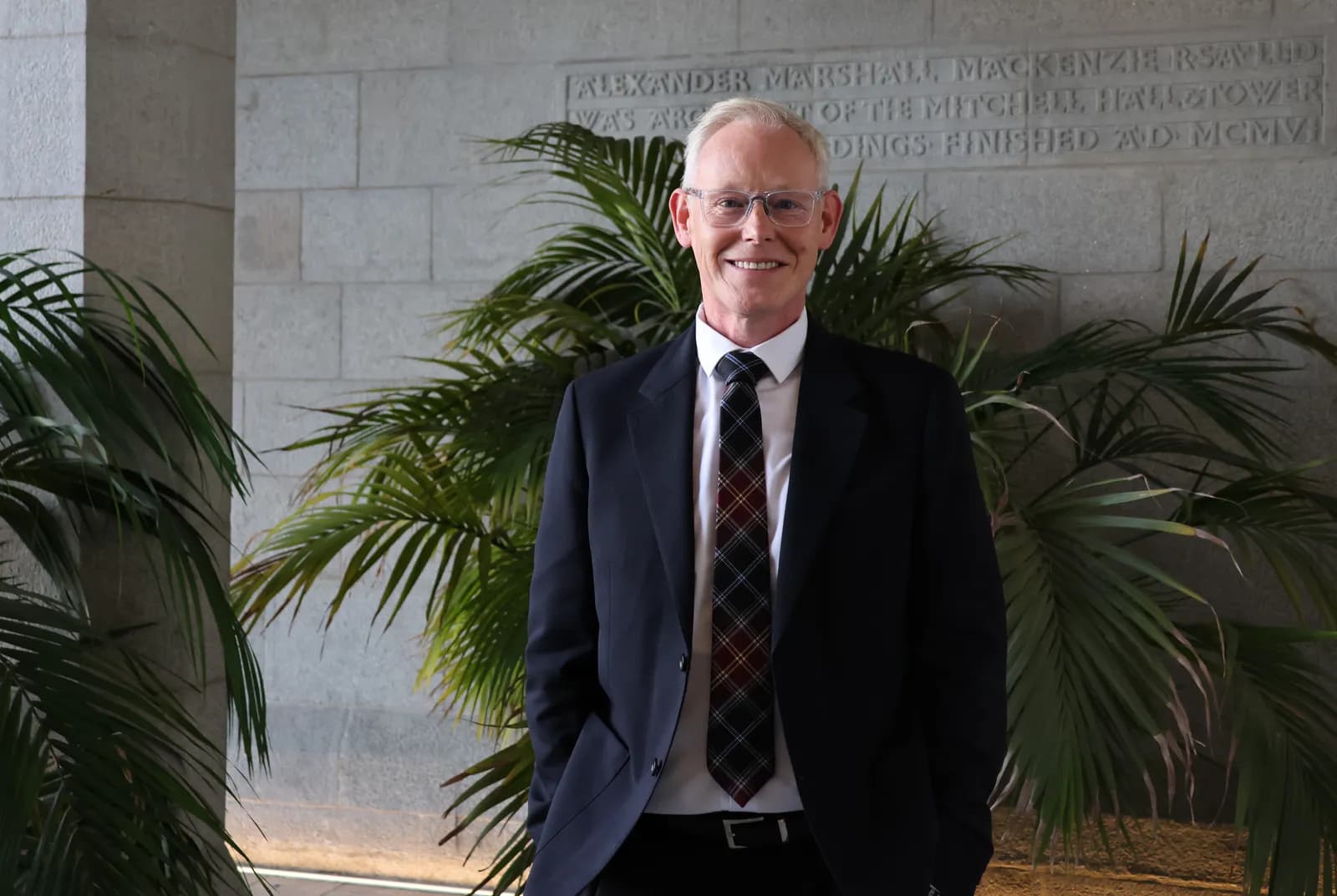A man in a suit and tie smiling with hands in pockets, standing in front of palm plants and a stone wall with engraved text.
