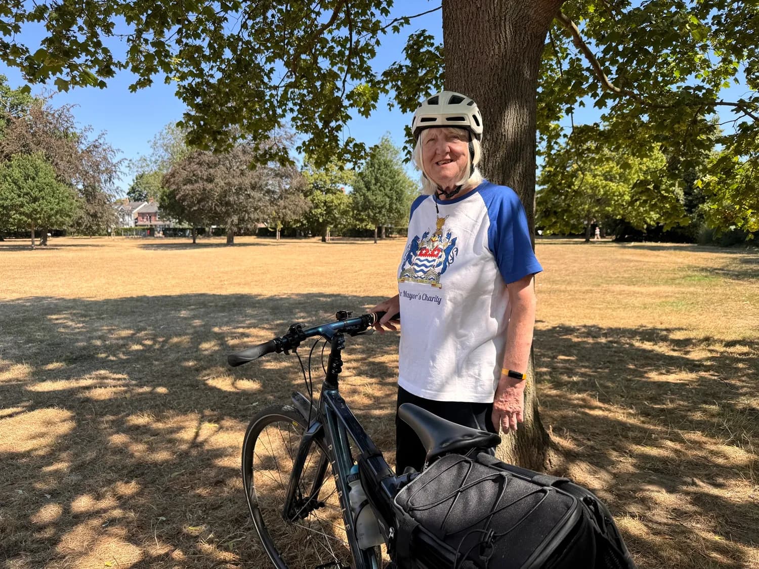 A woman wears a helmet and a white and blue shirt with a coat of arms logo while standing next to a bicycle under a tree in a sunny park with dry grass.