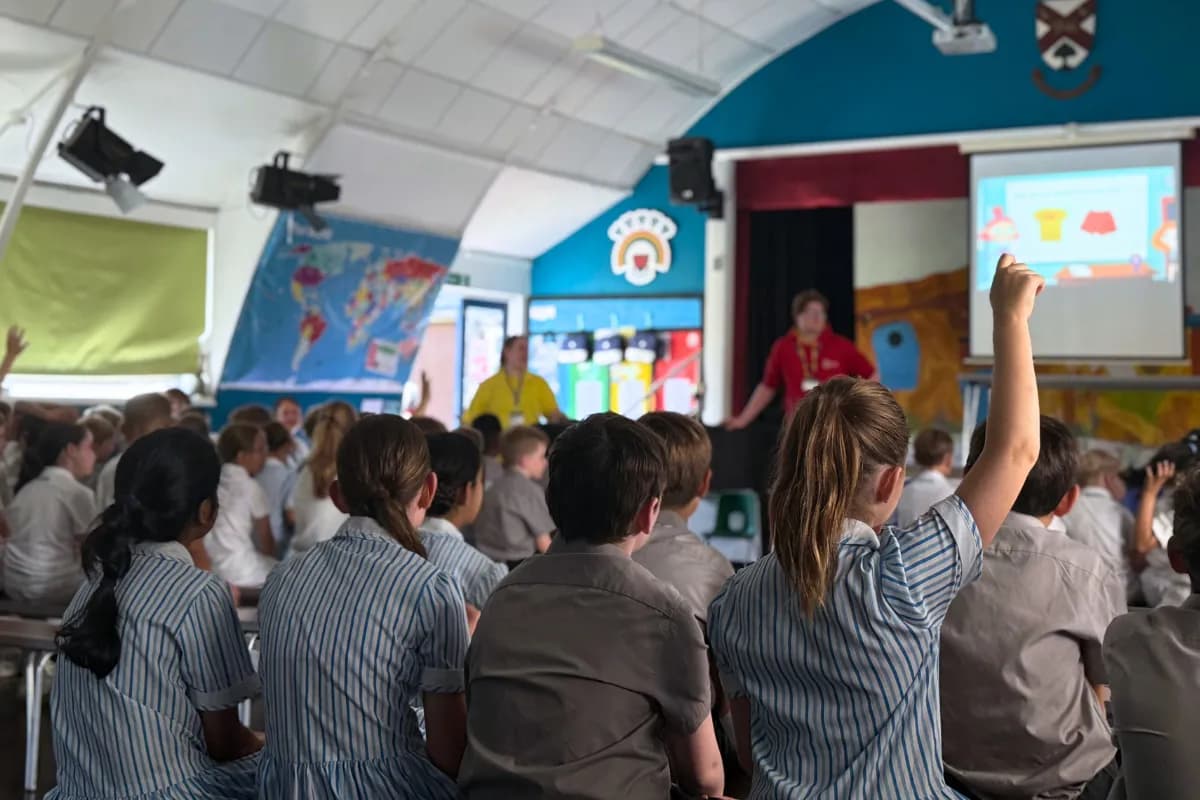 A group of schoolchildren sit in an assembly hall, with one student in the foreground raising her hand, while a presentation is projected onto a screen at the front.